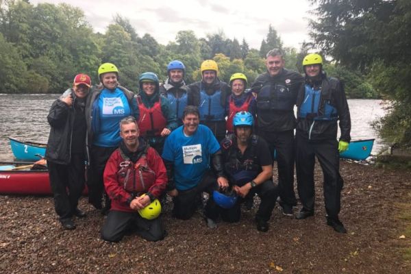 People in canoeing gear are ready for an expedition. They are beside a late surrounded by tress stood on the waters edge