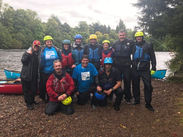 People in canoeing gear are ready for an expedition. They are beside a late surrounded by tress stood on the waters edge