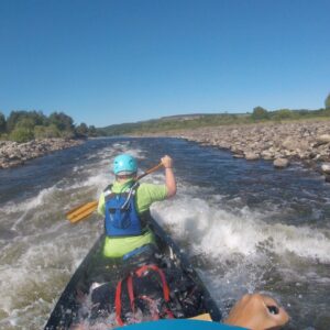 A person canoeing in some rapids on a river. He is wearing a helmet and life jacket whilst paddling away. The sky is bright blue and very sunny