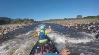 A person canoeing in some rapids on a river. He is wearing a helmet and life jacket whilst paddling away. The sky is bright blue and very sunny