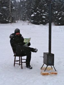 A man reading a piece of paper next to an outside log burner in snowy Sweden. He is wearing a big warm coat and sat on a wooden chair.