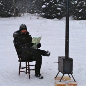 A man reading a piece of paper next to an outside log burner in snowy Sweden. He is wearing a big warm coat and sat on a wooden chair.