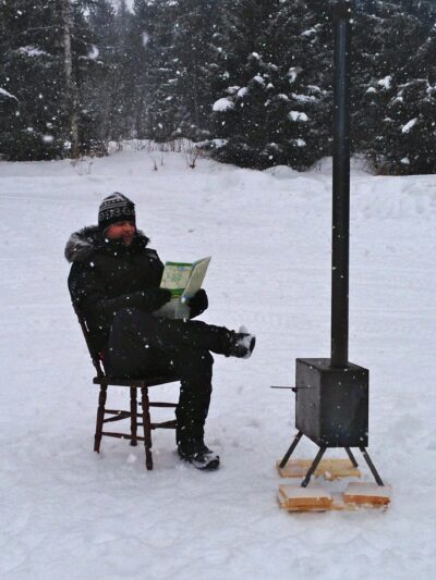 A man reading a piece of paper next to an outside log burner in snowy Sweden. He is wearing a big warm coat and sat on a wooden chair.