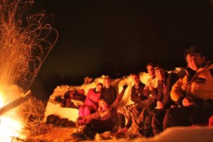A group of people on the Arctic-Style Survival Challenge huddled around a fire smiling at the camera.
