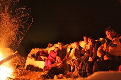 A group of people on the Arctic-Style Survival Challenge huddled around a fire smiling at the camera.