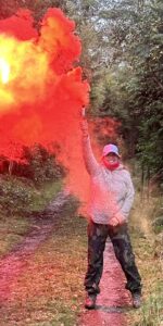 A lady stood in a forest with a red and orange smoke flare. She is smiling at the camera as she takes part in the UK Charity Survival Challenge.