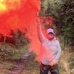 A lady stood in a forest with a red and orange smoke flare. She is smiling at the camera as she takes part in the UK Charity Survival Challenge.