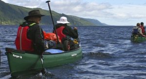 2 Canoes with 4 people between them. They are in green canoes paddling on the blue water. In the background, there is a mountain and a blue but moody sky.