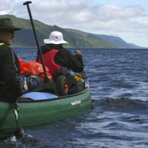2 Canoes with 4 people between them. They are in green canoes paddling on the blue water. In the background, there is a mountain and a blue but moody sky.