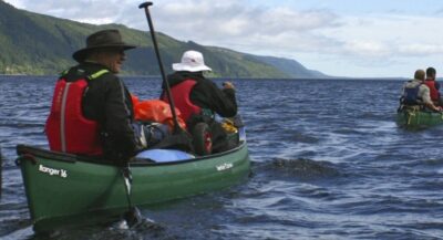 2 Canoes with 4 people between them. They are in green canoes paddling on the blue water. In the background, there is a mountain and a blue but moody sky.